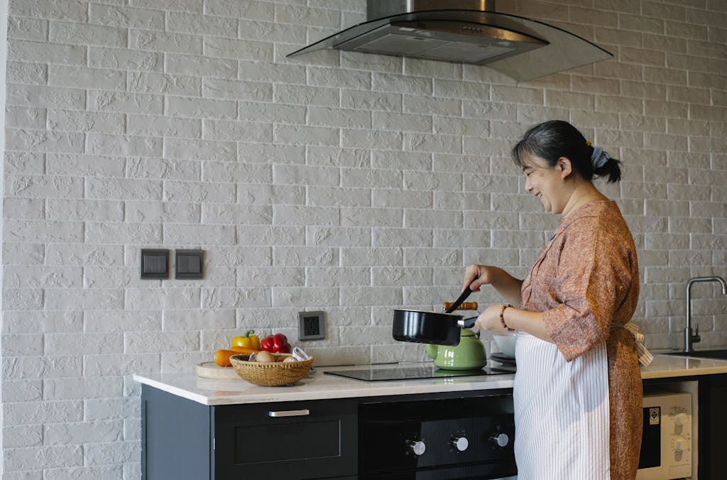 Side view of elderly female in apron standing near stove and holding pan while preparing lunch at home In a kitchen with range hood Kitchen exhaust fan